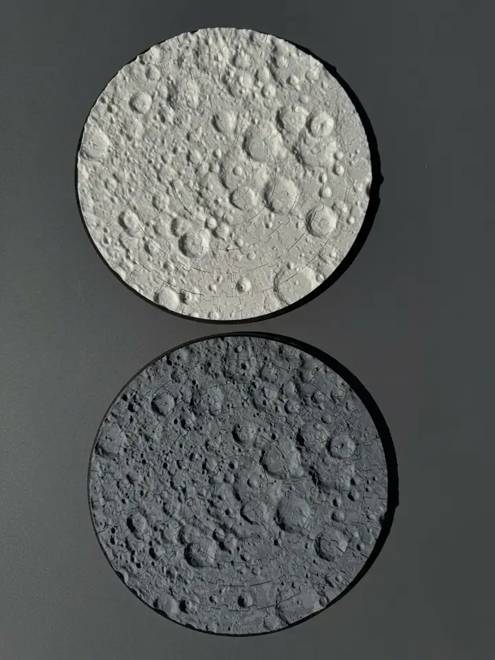Two finished Moon surface puzzles, one light and one dark, on a dark table.